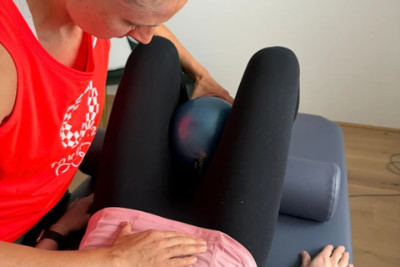 Übung bei Beckenbodenschwäche: liegende Person mit Ball zwischen den Beinen - unterstützt durch Physiotherapeutin im Beckenbereich