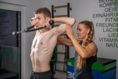Physiotherapeutin kontrolliert und korrigiert Bewegungsausführung eines Sportlers beim Training am Gerät
